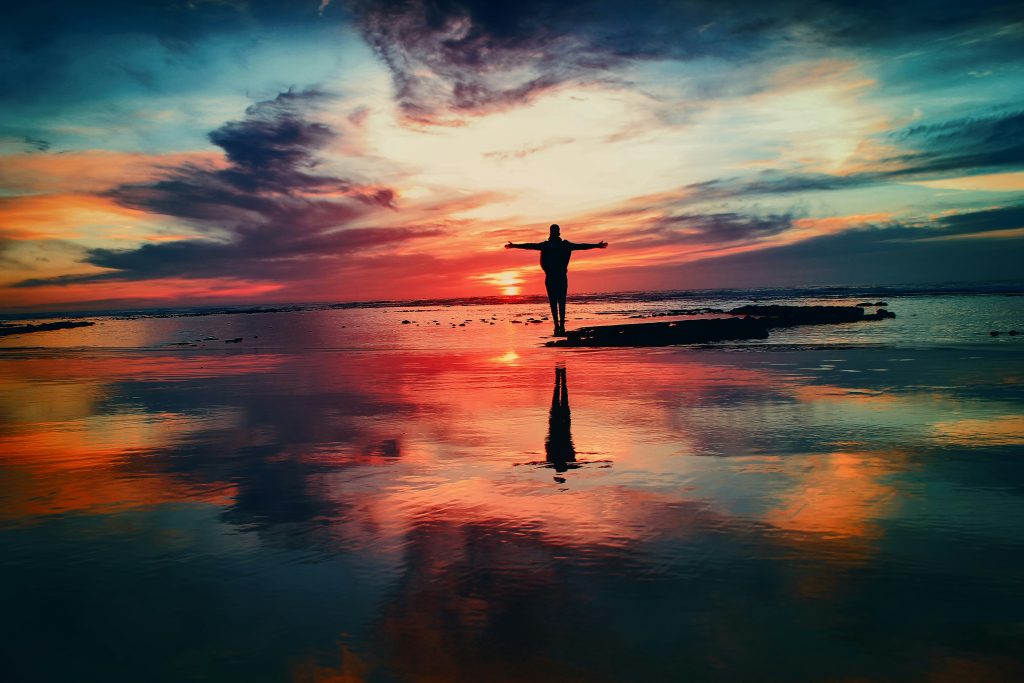 man standing on a beach at a colorful sunset with arms outstretched