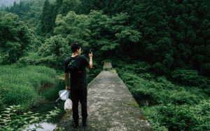 Asian man in black jeans and black tee-shirt holding an umbrella, with a dark backpack standing on a walkway taking a picture with a smart phone of the green trees and forest in front of him.
