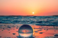 a clear glass ball sitting on the beach in the sand with an orange sunset glowing on the ball, the sand and the ocean