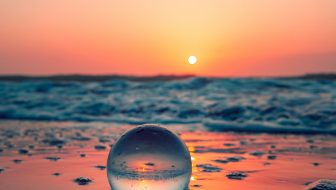 a clear glass ball sitting on the beach in the sand with an orange sunset glowing on the ball, the sand and the ocean